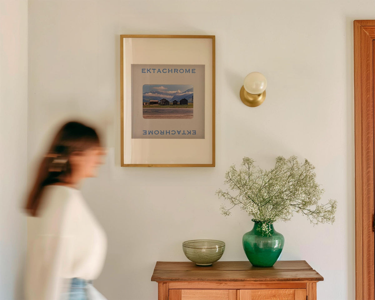 a woman walking past a framed picture on a wall, with a green vase with flowers and a bowl on a wooden table below the picture.