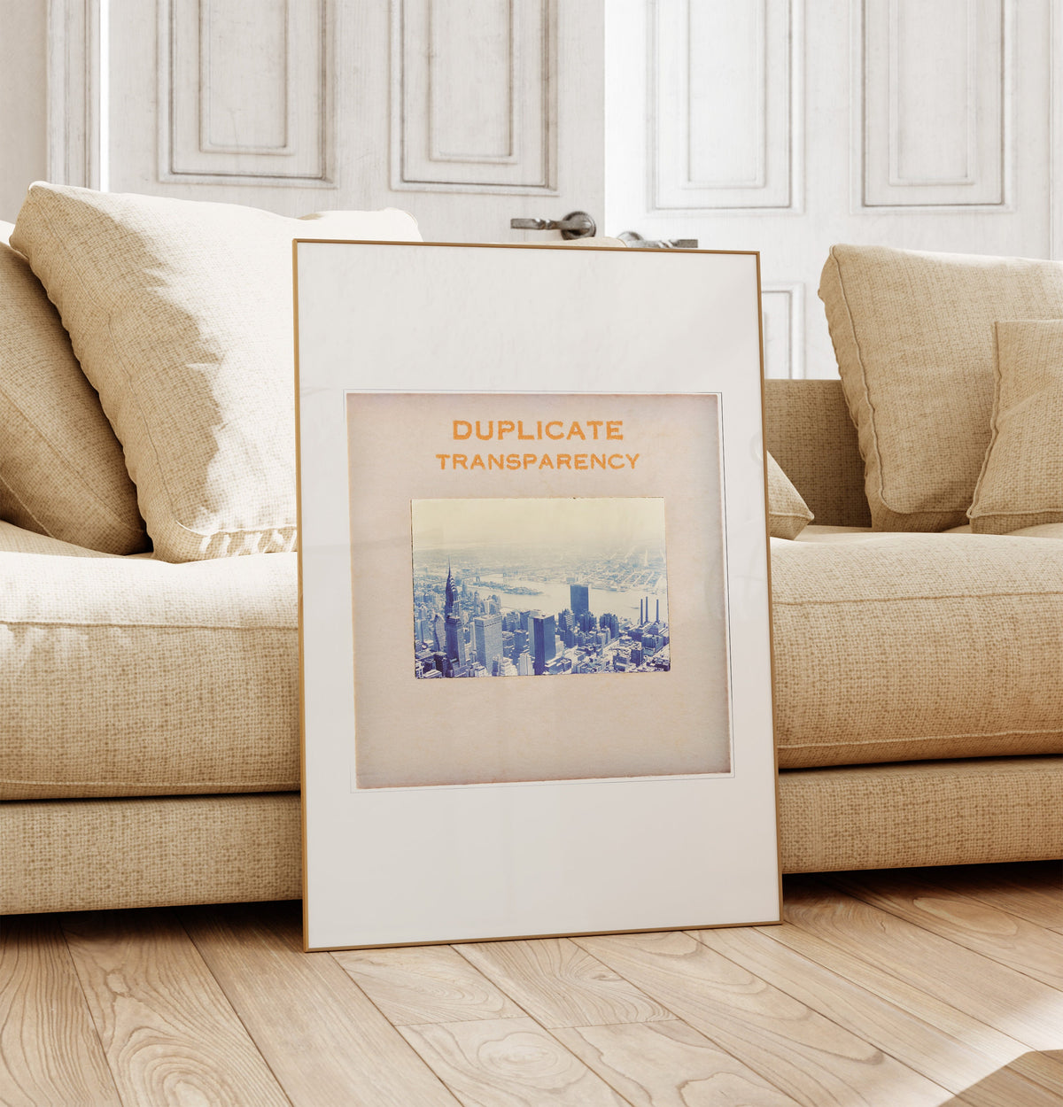 a framed picture of a cityscape, placed on a wooden floor in front of a beige sofa.
