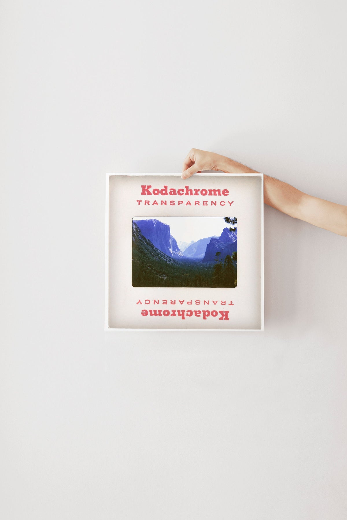 A person's hand is holding a framed photograph of a mountainous landscape, with the text "Kodachrome transparency" visible on the frame.