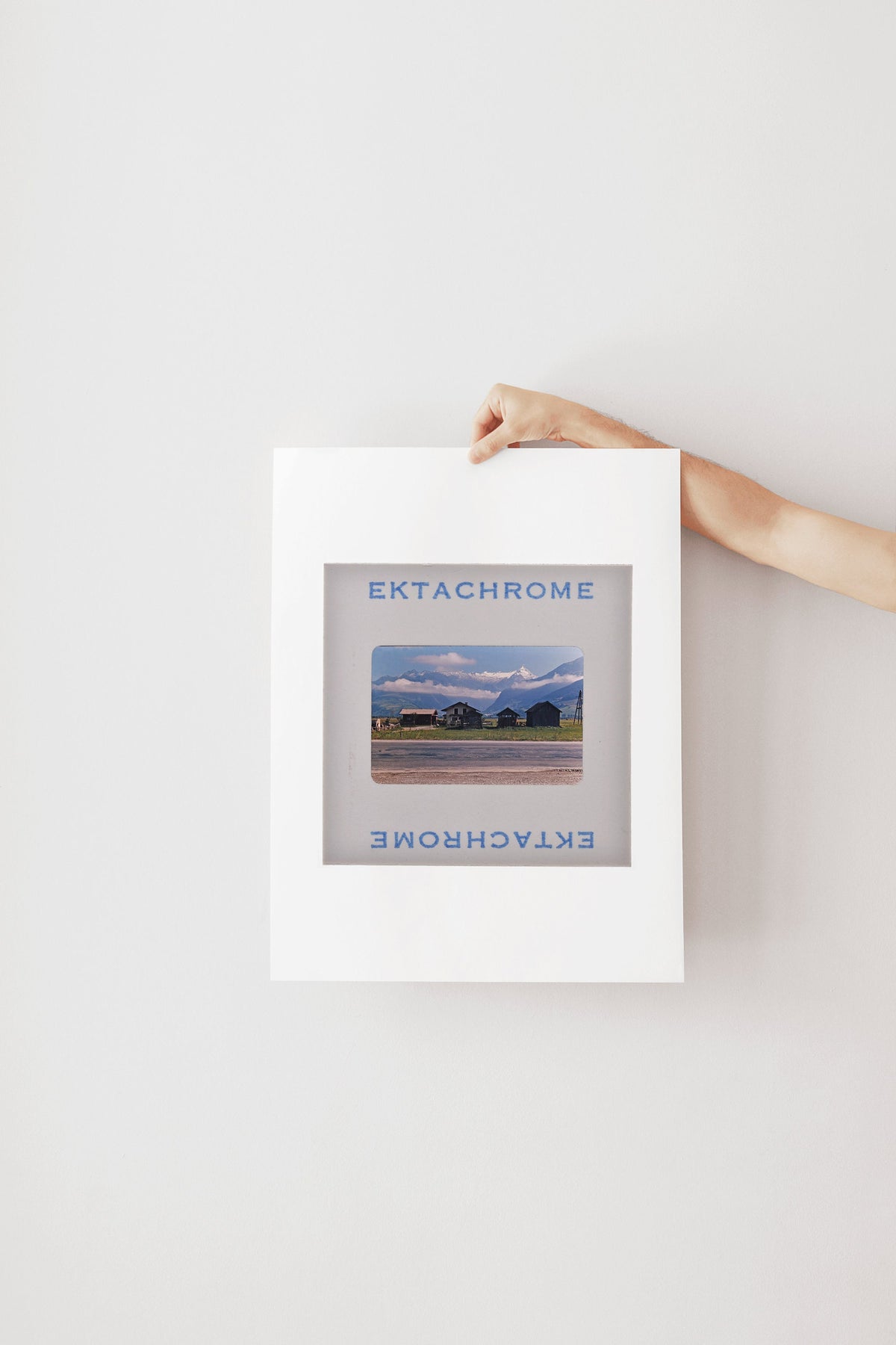 A person's hand is holding a framed photograph of a rural landscape, with the word "Ektachrome" written on the frame.