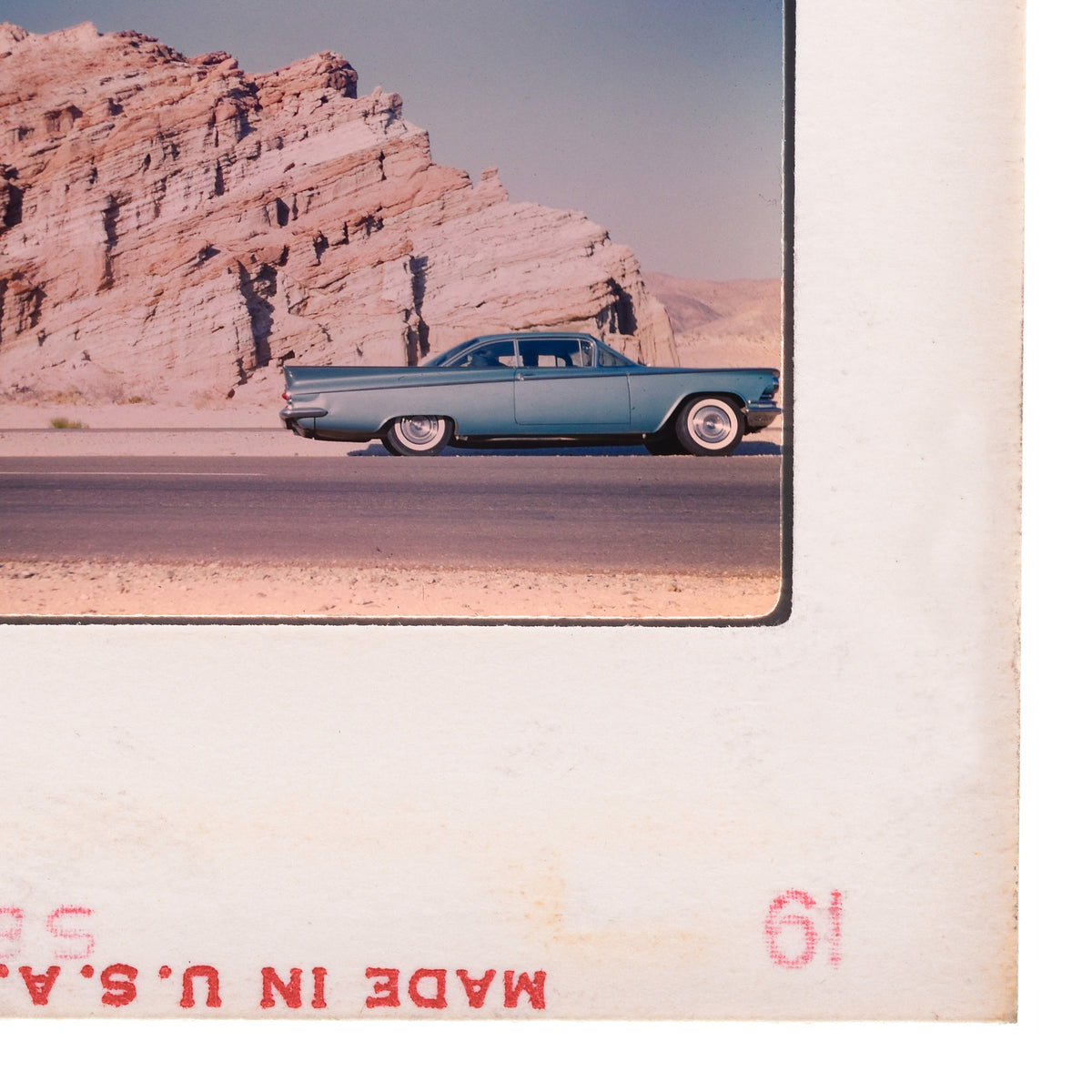 A vintage blue car is driving on a road in a desert landscape with a rocky cliff in the background.