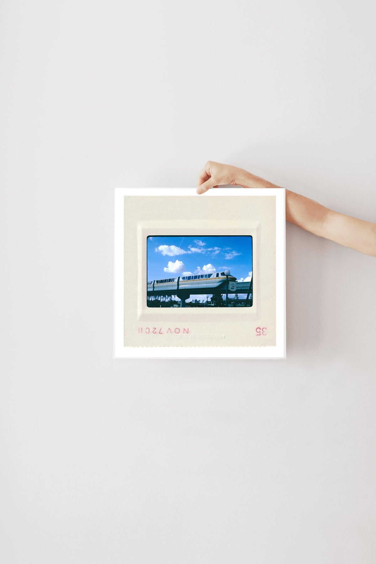 A person's hand is holding a framed picture of a train against a white wall.