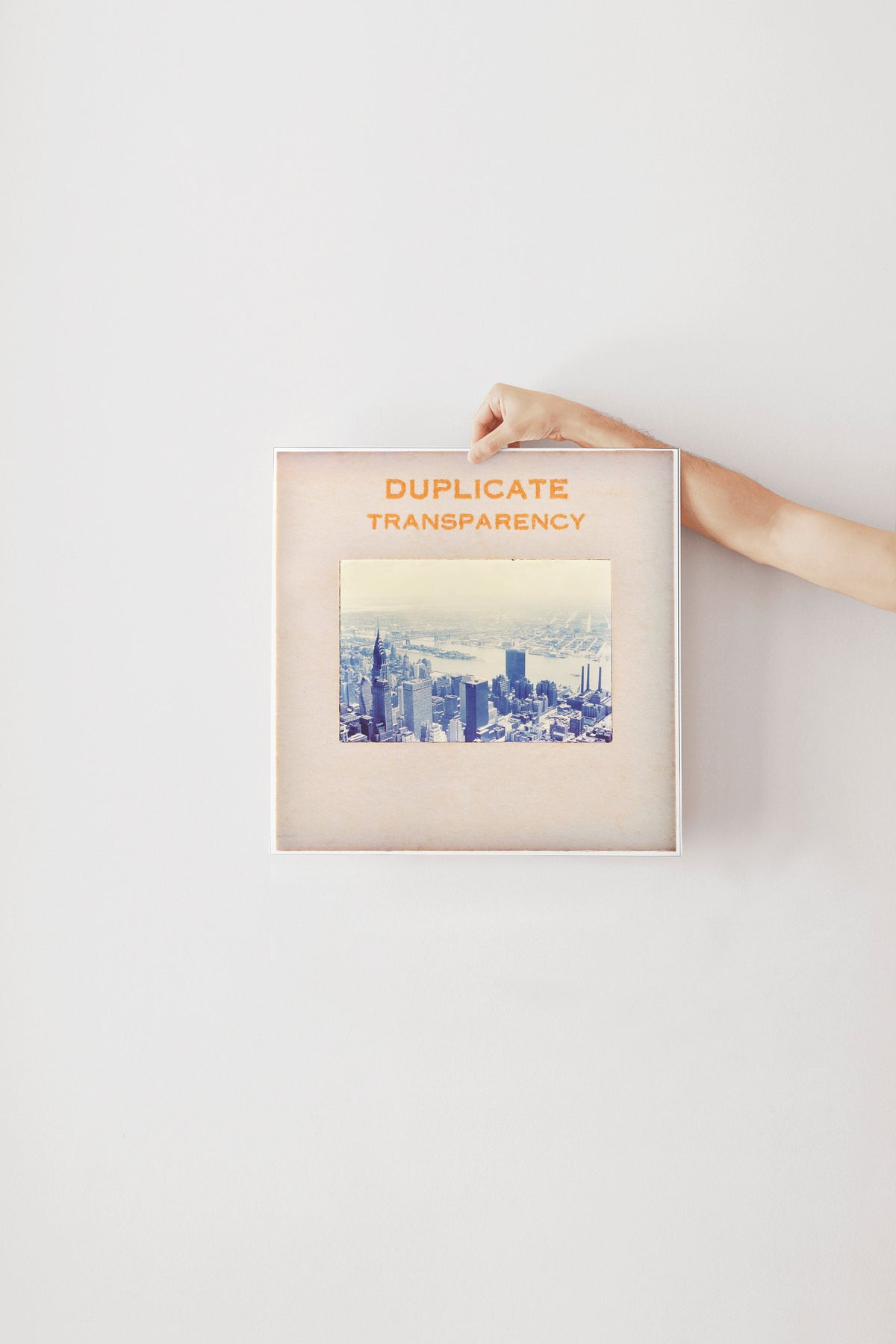 A person's hand is holding a framed picture of a cityscape, with the text "DUPLICATE TRANSPARENCY" written on the frame.