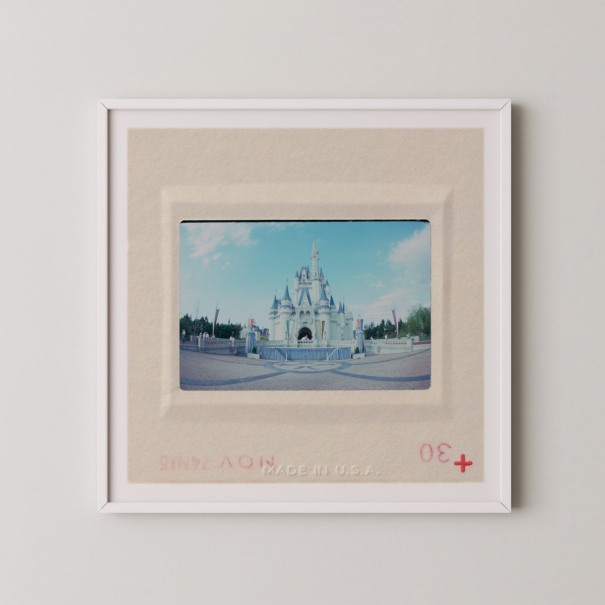 A framed photograph of a castle, likely from a theme park or tourist attraction, with a red border and a red plus sign in the bottom right corner.