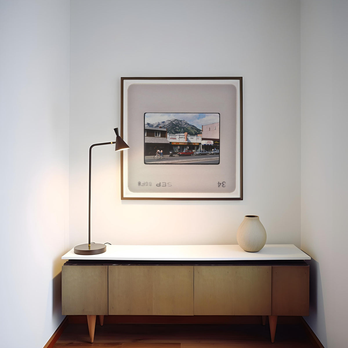 a minimalist corner of a room with a wooden cabinet, a lamp, and a framed picture of a building on the wall.