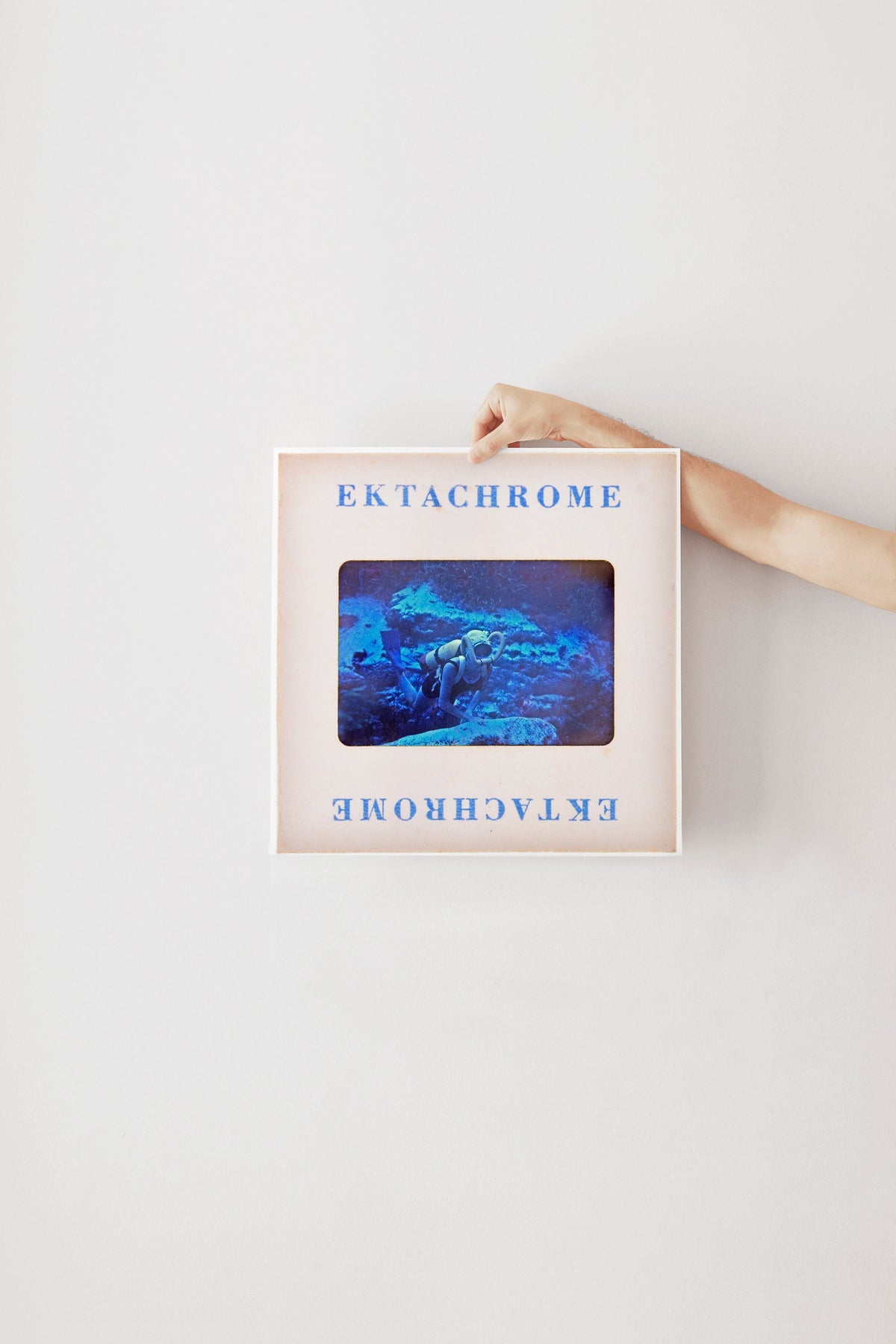 A person's hand is holding a framed image of a blue underwater scene, with the text "Ektachrome" visible on the frame.