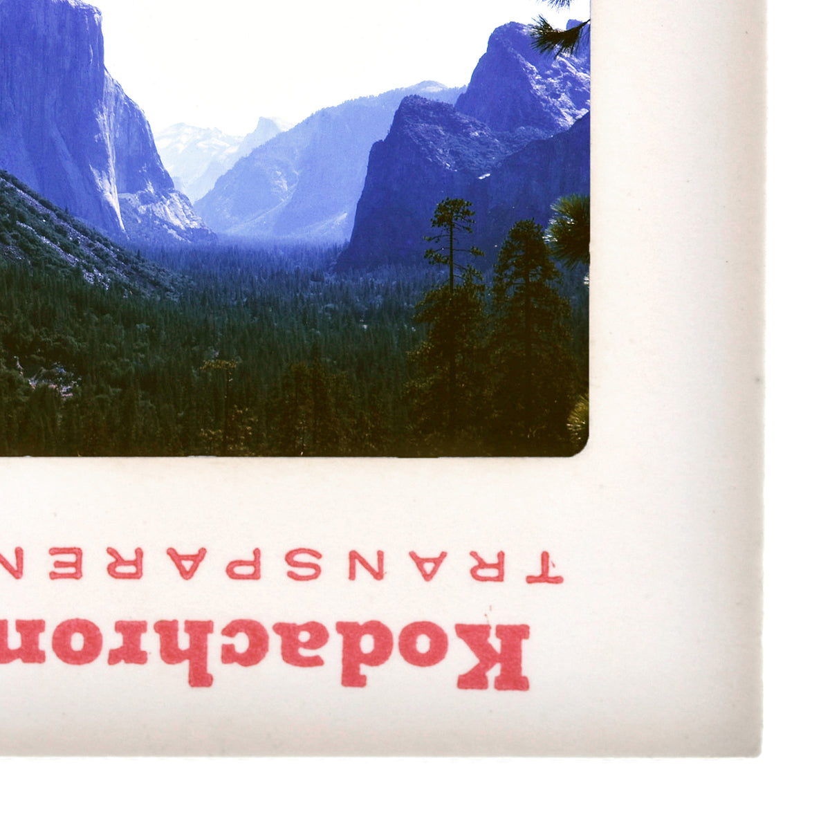 A framed photograph of a mountainous landscape with trees and snow-capped peaks, featuring a red and white text overlay that reads "Kodak".