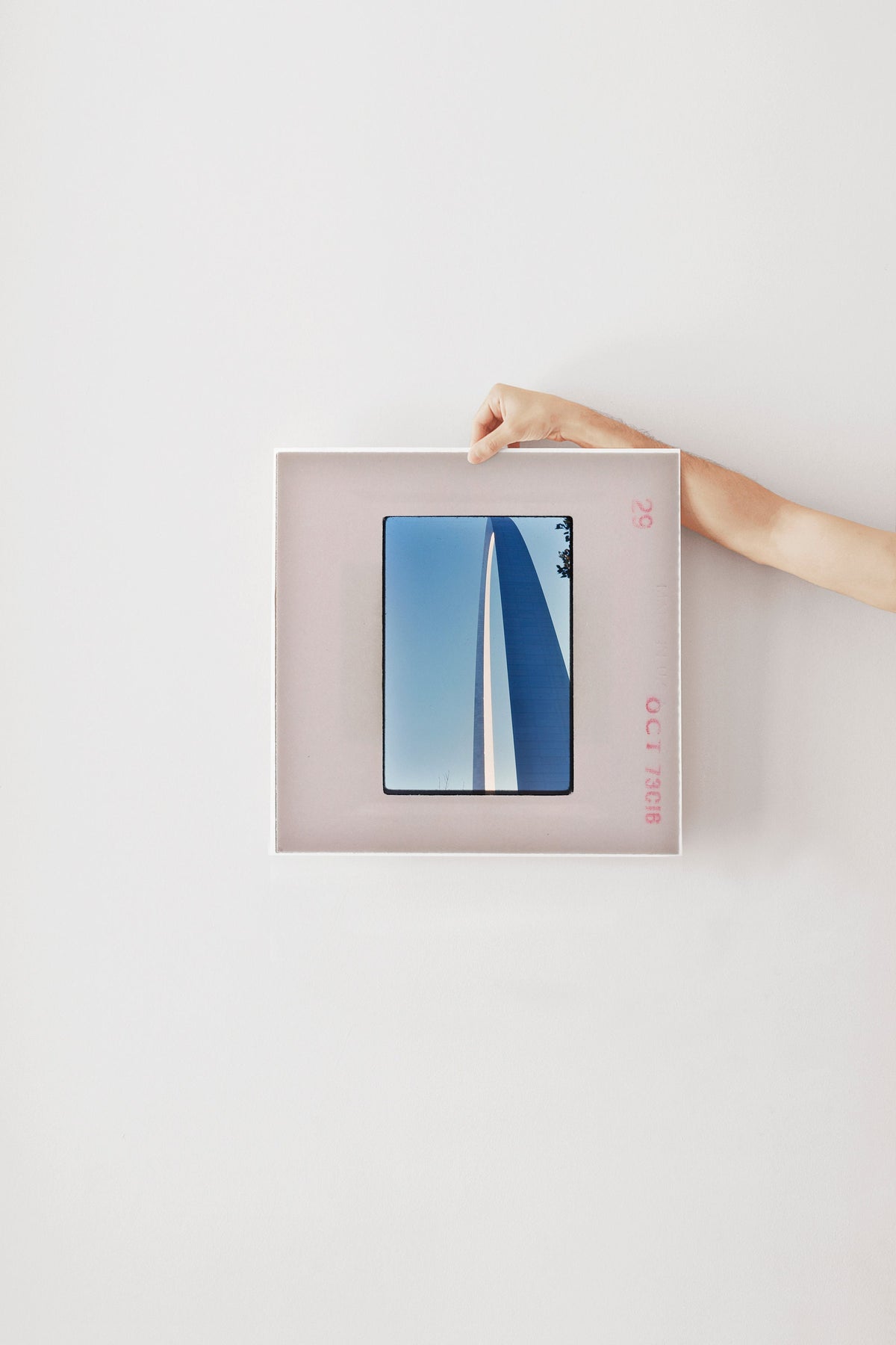 A person's hand is holding a framed photograph of a blue sky with a white stripe, against a plain white wall.