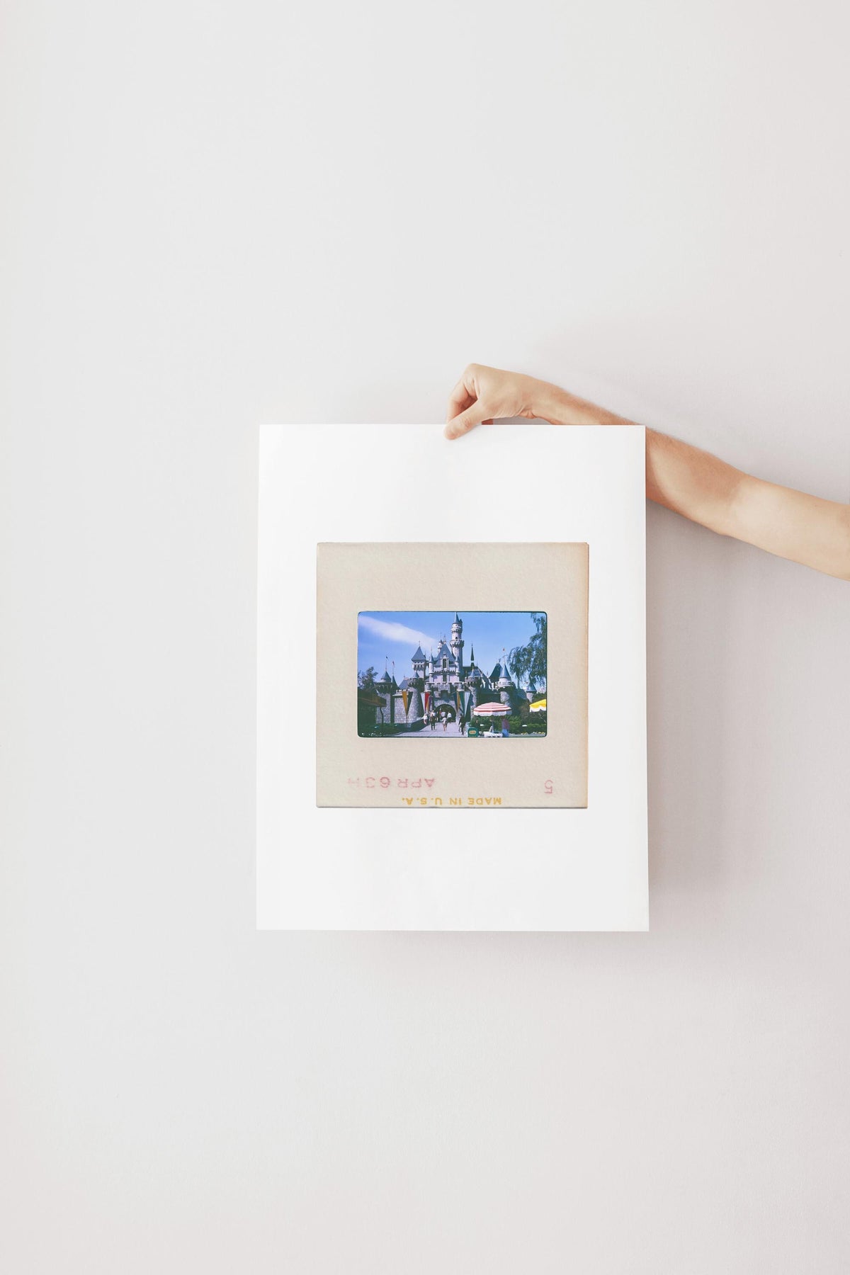 A person's hand is holding a framed picture of a castle or palace, which is hanging on a white wall.