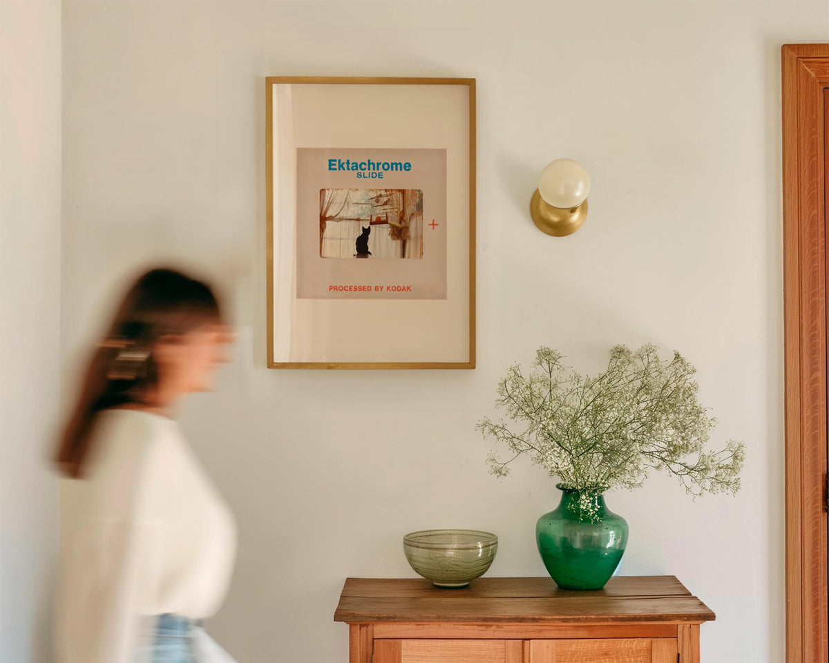 a woman walking past a wooden table with a green vase and a framed picture on the wall.