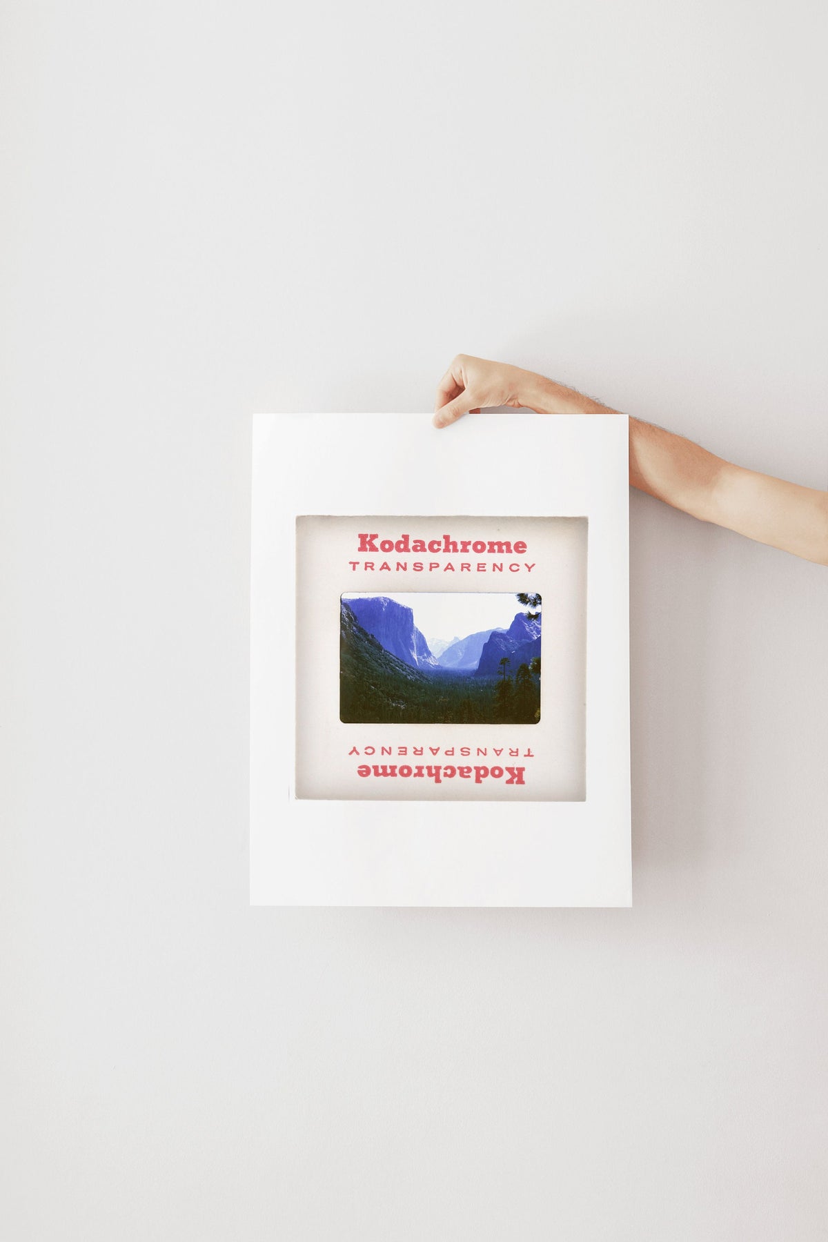 A hand is holding a framed photograph of a mountainous landscape, with the text "Kodachrome Transparency" visible on the frame.