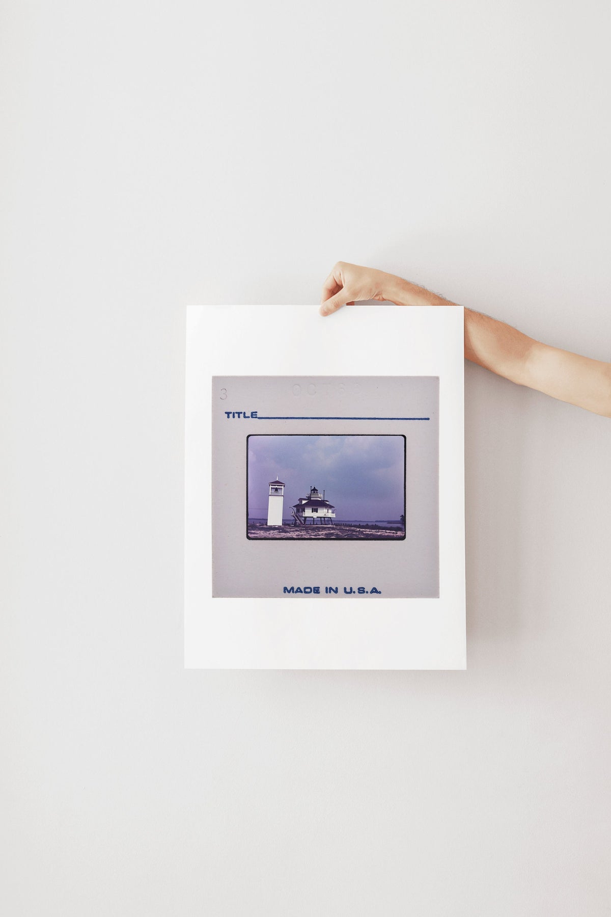 A person's hand is holding a framed photograph of a lighthouse against a white background.