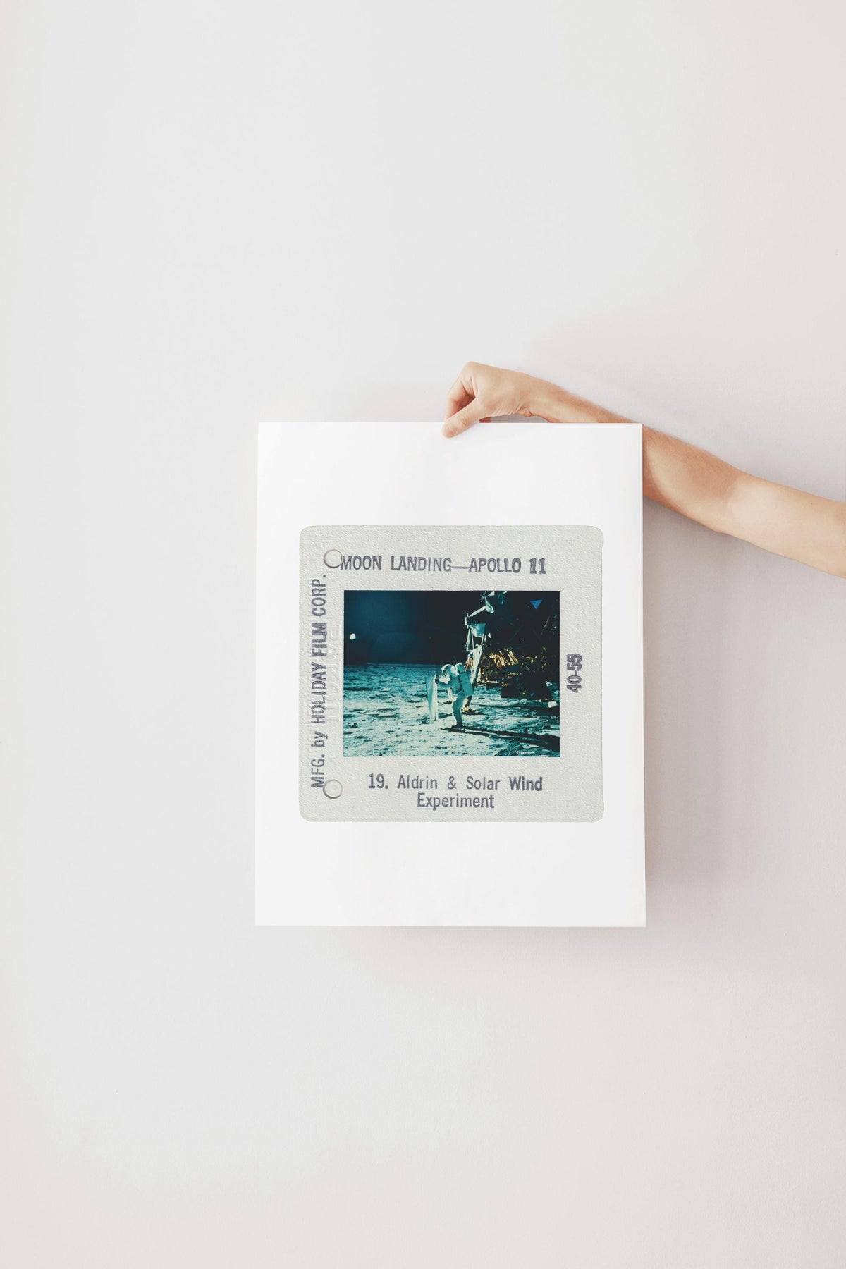 A person's hand is holding a framed photograph of the Apollo 11 moon landing, with the text "Moon Landing - Apollo 11" and "18. Aldrin & Solar Wind Experiment" visible.