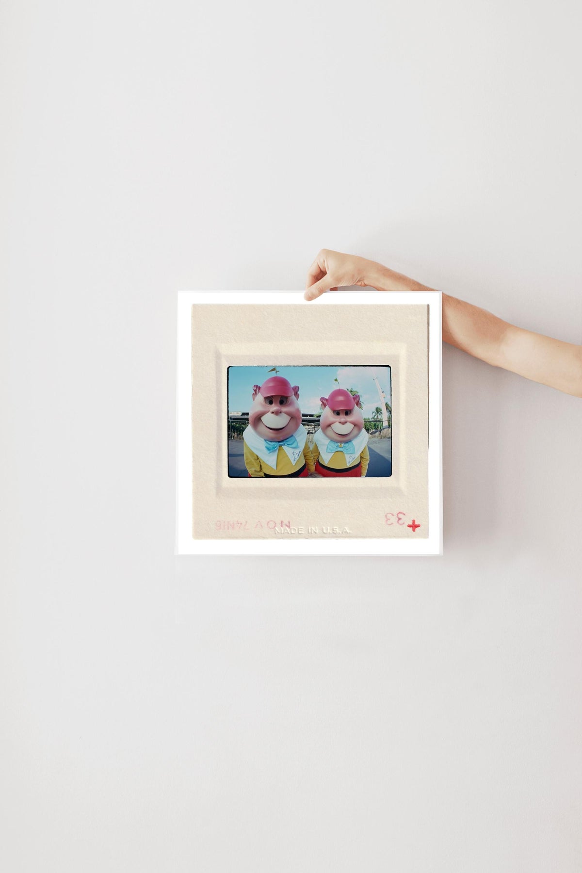 A person's hand is holding a white picture frame with a framed image of two cartoon characters, likely a pair of anthropomorphic animals, on the wall.