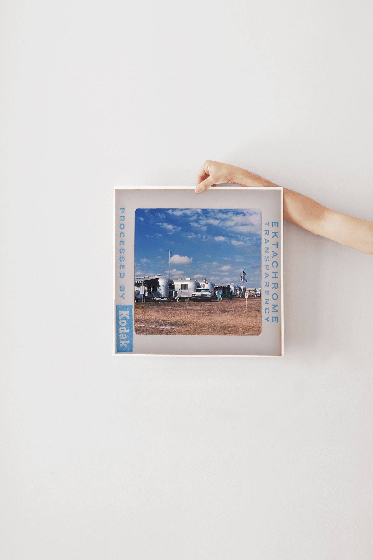 A hand is holding a framed photograph of a beach scene with a camper trailer in the background.