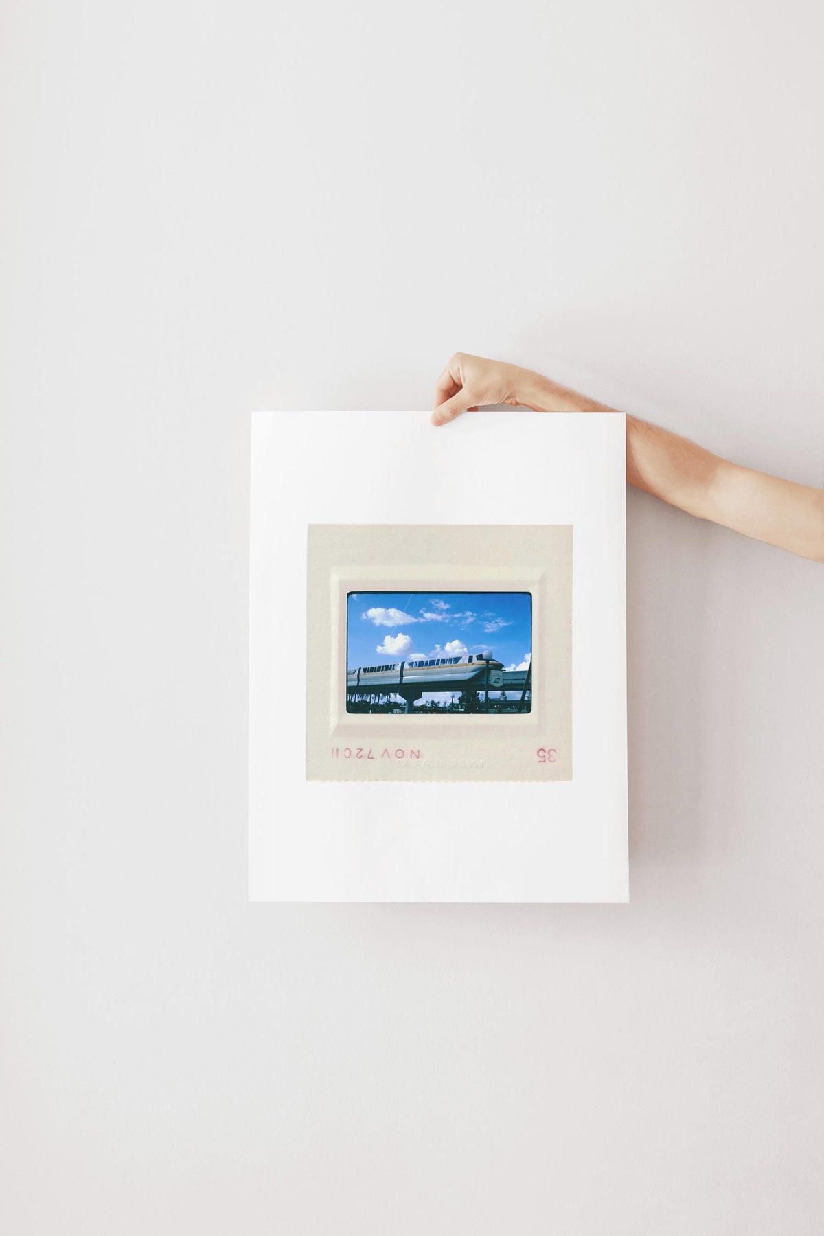 A person's hand is holding a framed picture of a train against a white wall.