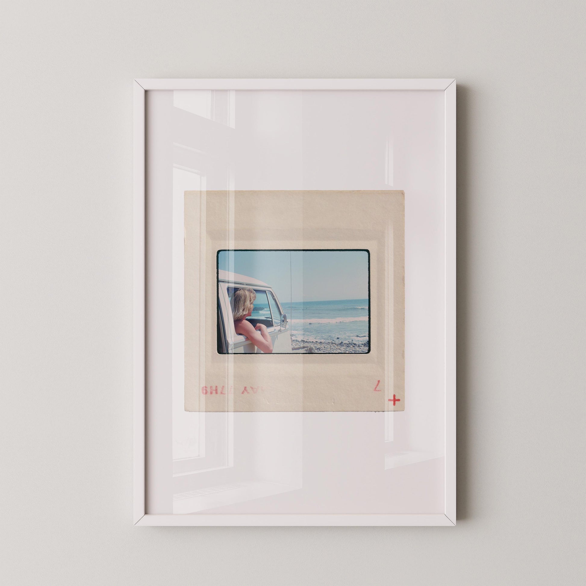 Vintage 1977 Kodachrome slide photograph of a blonde child leaning out the window of a white Volkswagen Microbus, looking toward the ocean along the California coast. Rocky shoreline and gentle surf visible in the background. Printed as a giclée fine art print on archival matte paper.