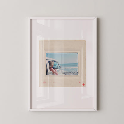 Vintage 1977 Kodachrome slide photograph of a blonde child leaning out the window of a white Volkswagen Microbus, looking toward the ocean along the California coast. Rocky shoreline and gentle surf visible in the background. Printed as a giclée fine art print on archival matte paper.