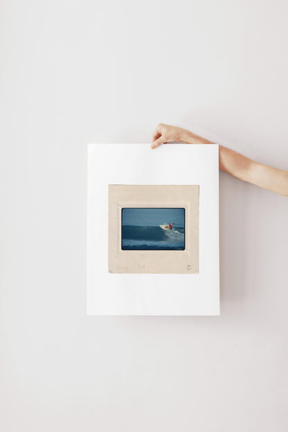 Vintage 1970s Kodachrome slide photograph of a surfer in a red and yellow competition jersey carving off the top of a deep blue wave along the California coast, with white spray exploding from the board. Printed as a giclée fine art print on archival matte paper.