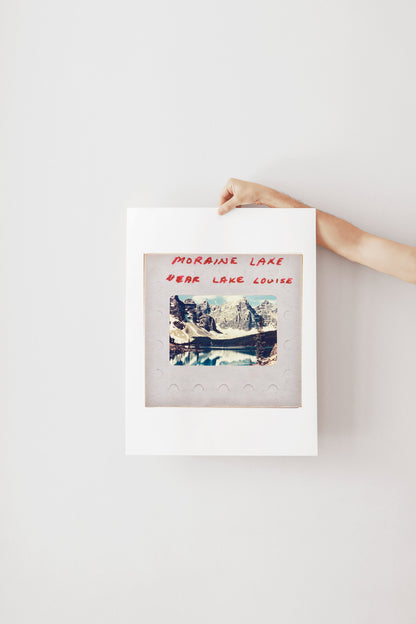 A hand is holding a framed photograph of a scenic mountain landscape, with the text "MORaine Lake" and "EAR Lake Louise" written on the frame.