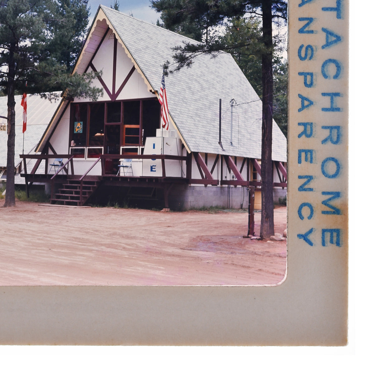 A white wooden cabin with a triangular roof, surrounded by trees, with a flagpole and a sign on the front of the building.