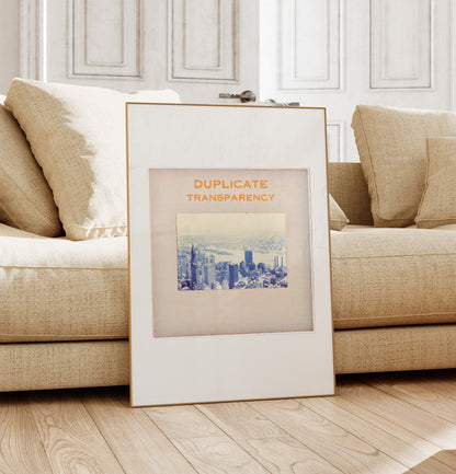 a framed picture of a cityscape, placed on a wooden floor in front of a beige sofa.