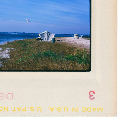 A white tent is set up on a grassy beach, with a bird flying overhead and a van parked nearby.