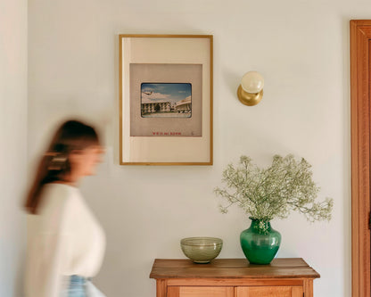 a woman walking past a wooden table with a framed picture, a green vase with flowers, and a bowl on it. The table is located in a hallway, and the woman is blurred, indicating movement.