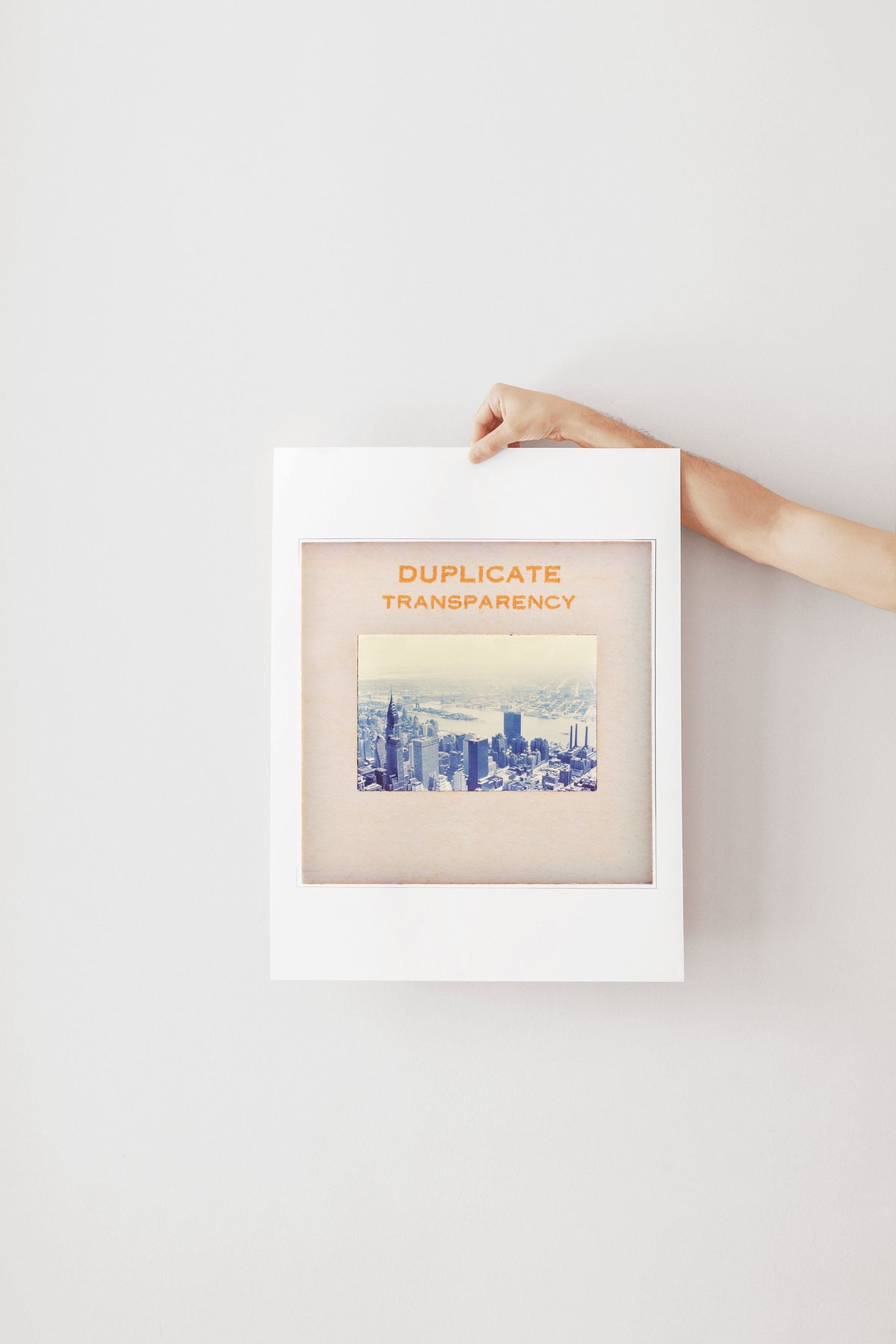 A person's hand is holding a framed picture of a cityscape, with the text "DUPLICATE TRANSPARENCY" written on the frame.