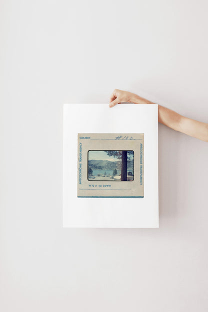 Vintage Anscochrome slide photograph of a mountain lake campground community, showing pine-forested hills, a snow-capped peak, classic cars parked along the shoreline, and lakeside cabins, viewed from beneath a tall pine tree. Printed as a giclée fine art print on archival matte paper.