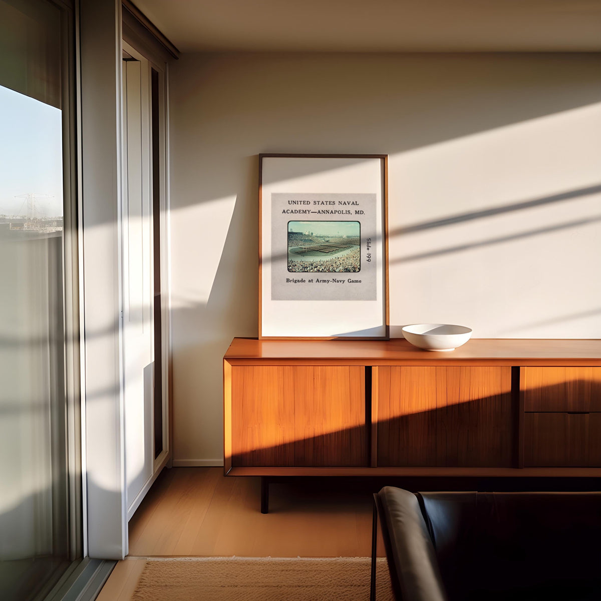 a wooden cabinet with a white bowl on top, situated in a room with a large window and a framed poster of a landscape on the wall.