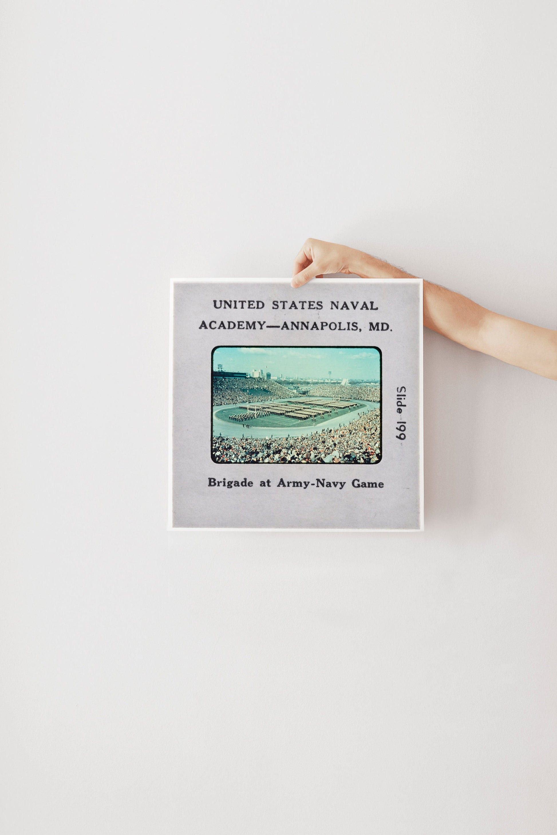 A person's hand is holding a framed photograph of a naval game, with text indicating it is from the United States Naval Academy in Annapolis, MD.