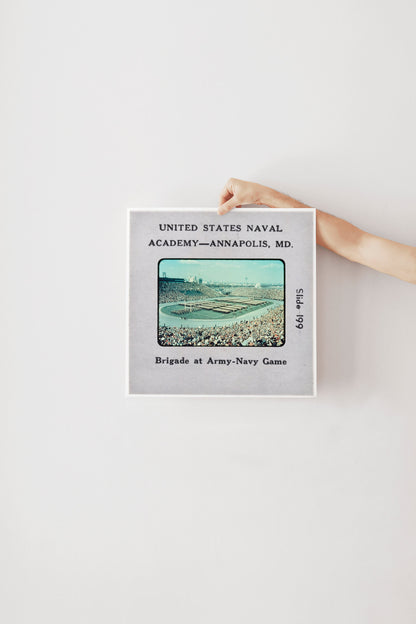 A person's hand is holding a framed photograph of a naval game, with text indicating it is from the United States Naval Academy in Annapolis, MD.