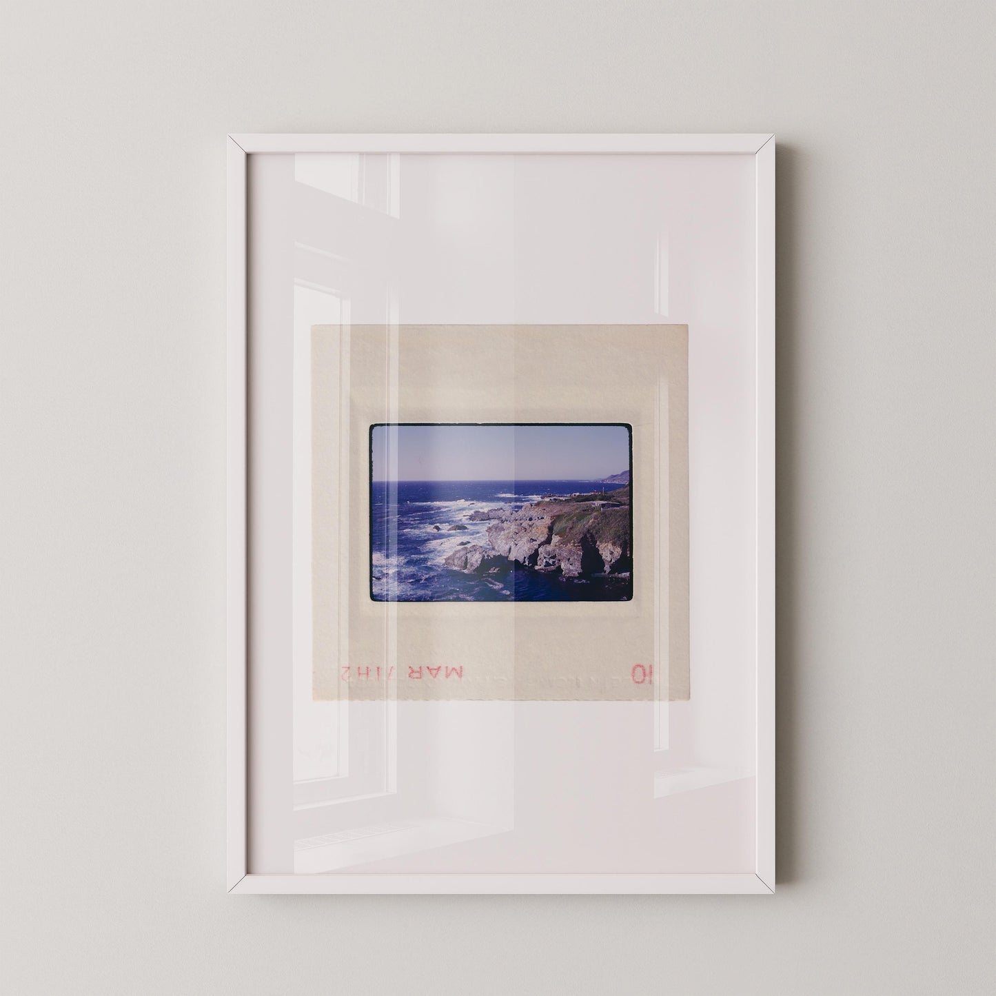 Vintage 1971 Kodachrome slide photograph of the Carmel California coastline, showing dramatic rocky cliffs and outcroppings meeting deep cobalt Pacific Ocean waters, with white surf, a green headland, and a small structure visible at the cliff's edge. Printed as a giclée fine art print on archival matte paper.