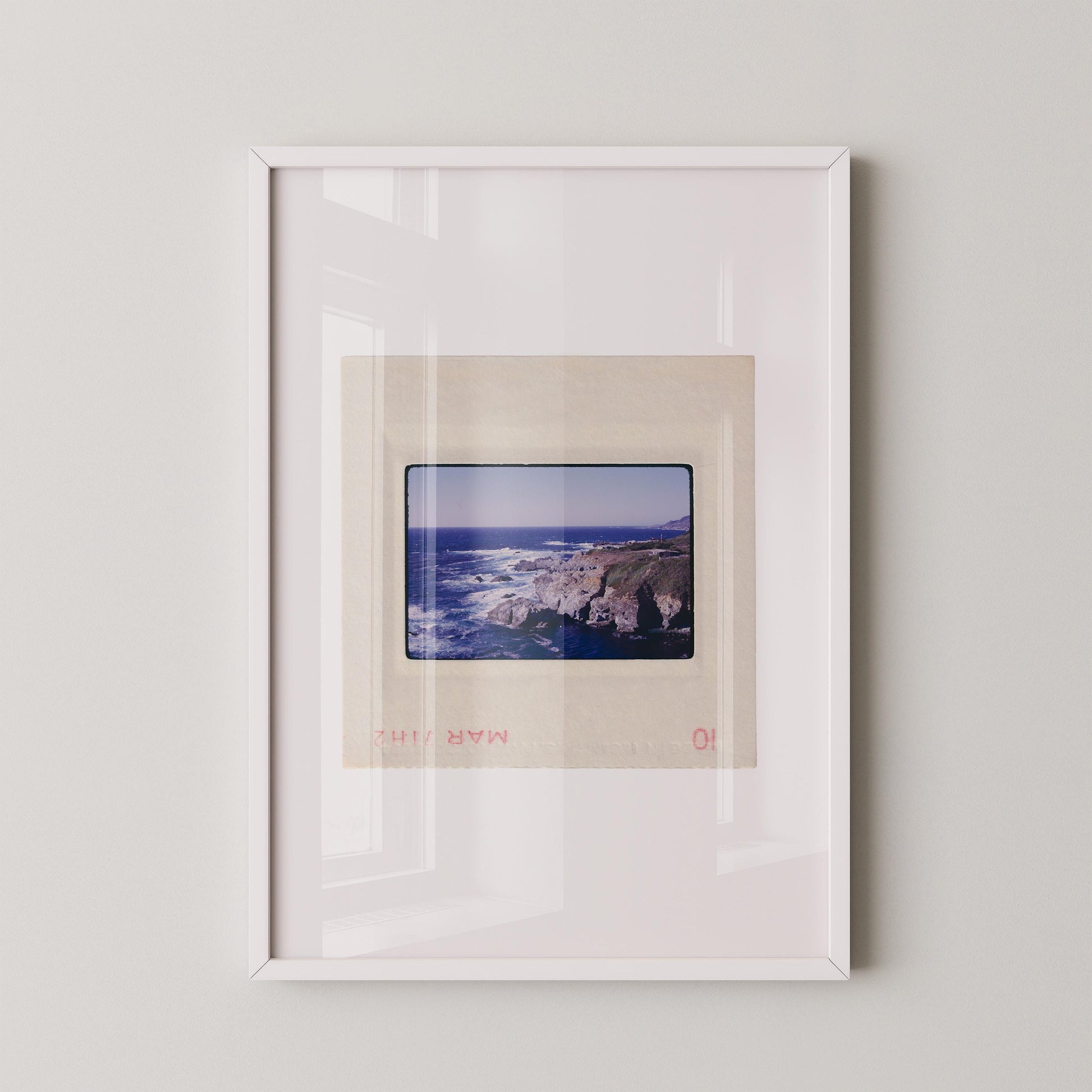 Vintage 1971 Kodachrome slide photograph of the Carmel California coastline, showing dramatic rocky cliffs and outcroppings meeting deep cobalt Pacific Ocean waters, with white surf, a green headland, and a small structure visible at the cliff's edge. Printed as a giclée fine art print on archival matte paper.