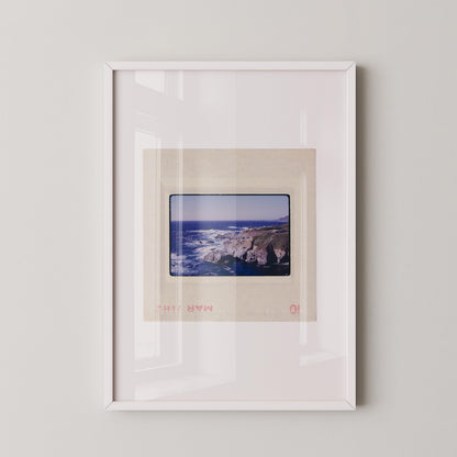 Vintage 1971 Kodachrome slide photograph of the Carmel California coastline, showing dramatic rocky cliffs and outcroppings meeting deep cobalt Pacific Ocean waters, with white surf, a green headland, and a small structure visible at the cliff's edge. Printed as a giclée fine art print on archival matte paper.