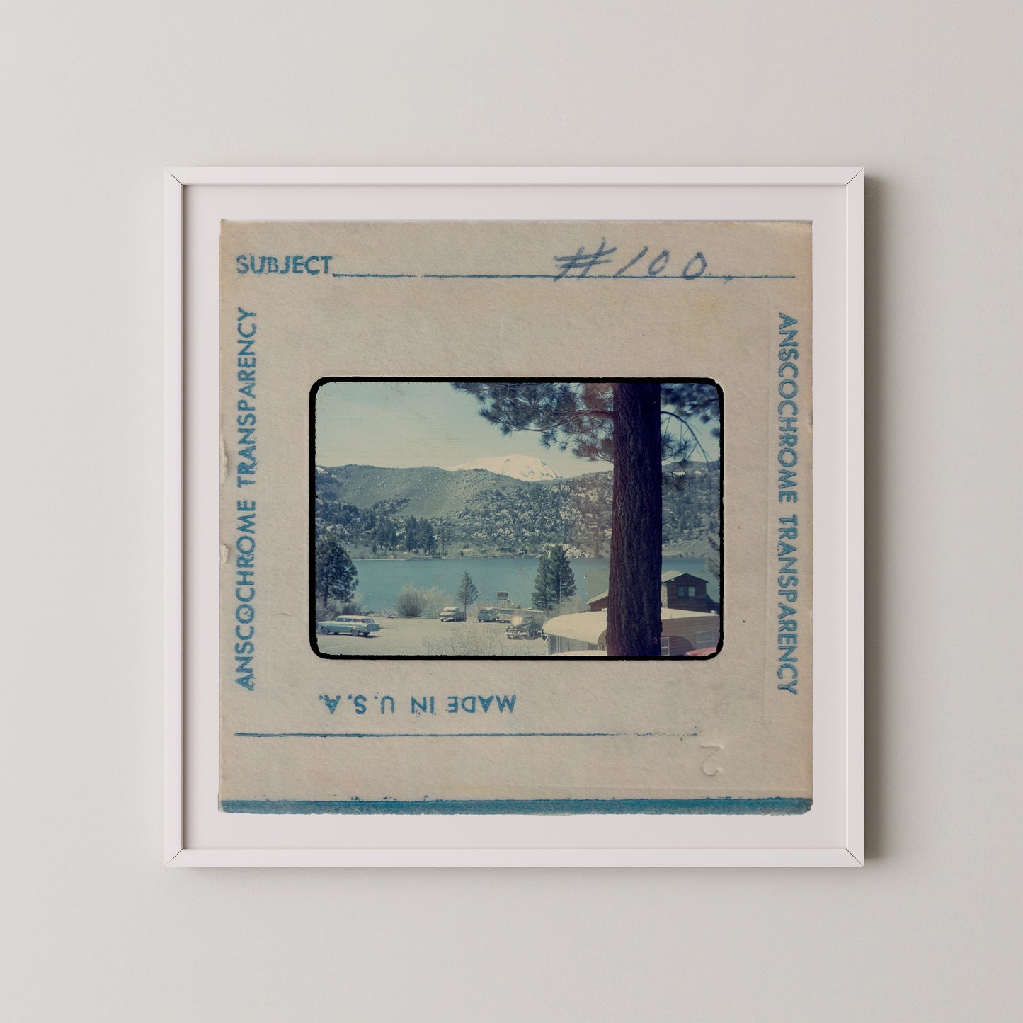 The image is a framed photograph of a scenic landscape, featuring a lake surrounded by mountains and trees.