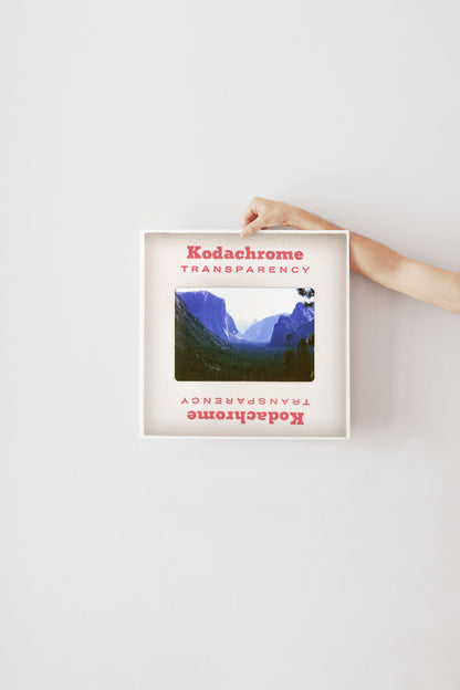 A person's hand is holding a framed photograph of a mountainous landscape, with the text "Kodachrome transparency" visible on the frame.