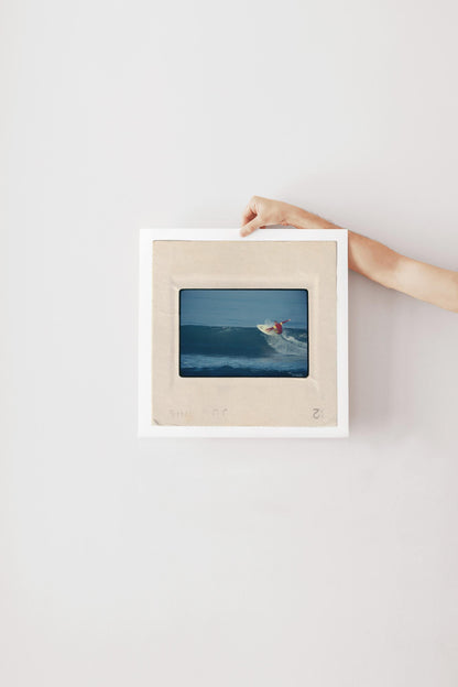Vintage 1970s Kodachrome slide photograph of a surfer in a red and yellow competition jersey carving off the top of a deep blue wave along the California coast, with white spray exploding from the board. Printed as a giclée fine art print on archival matte paper.