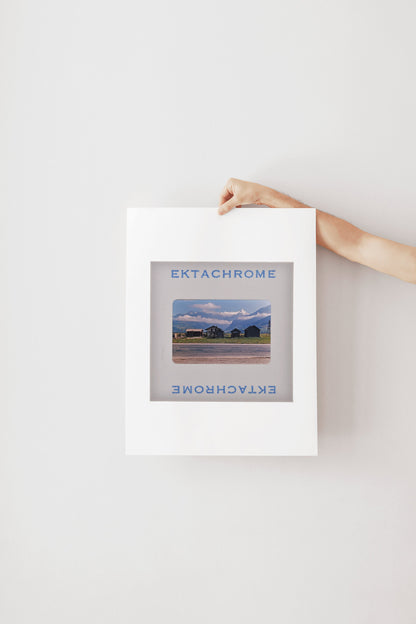 A person's hand is holding a framed photograph of a rural landscape, with the word "Ektachrome" written on the frame.