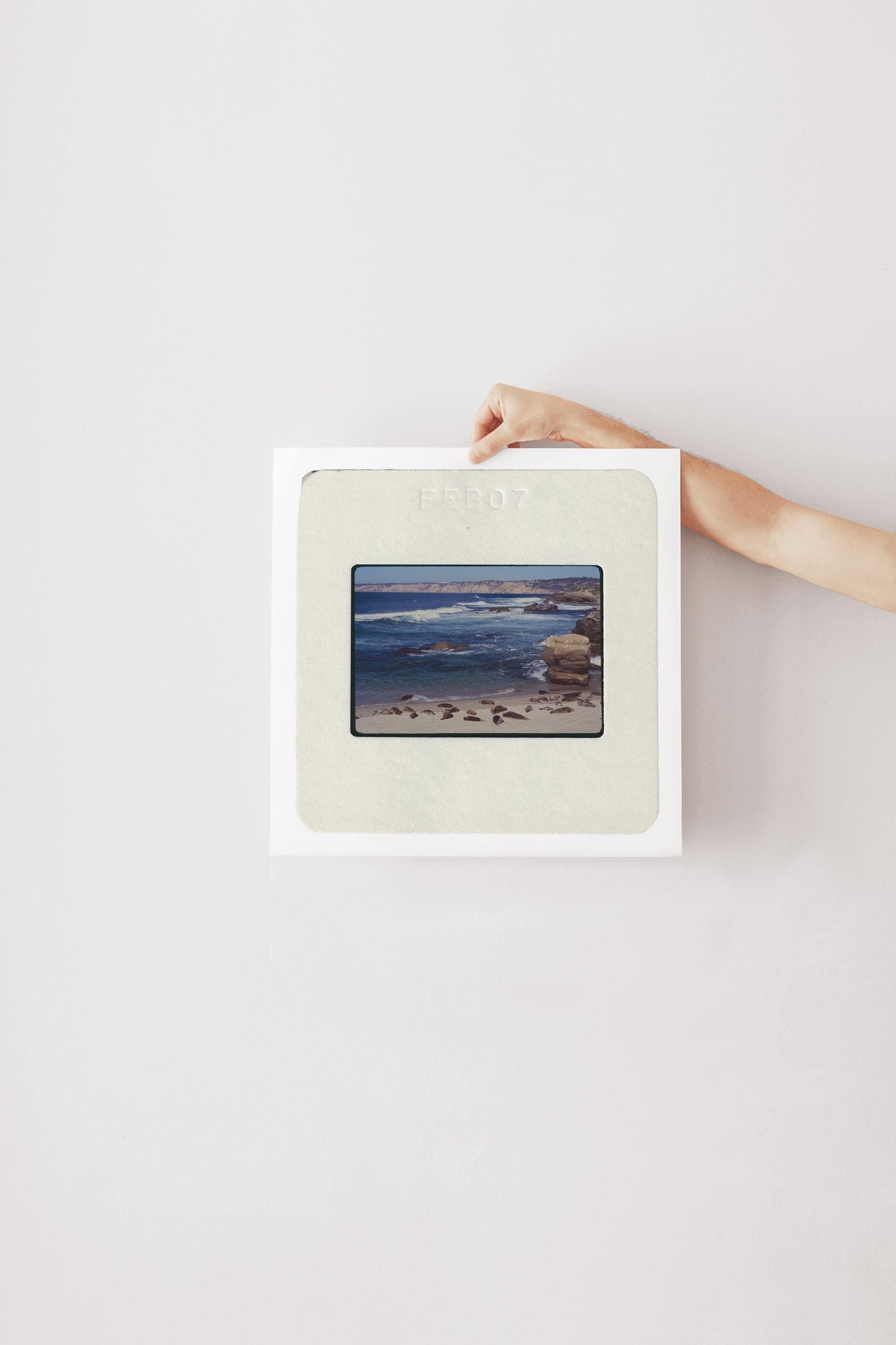 Vintage color slide photograph of dozens of harbor seals resting on a sandy California beach, with dramatic sandstone rock formations, rolling Pacific surf, and coastal bluffs visible in the background, likely La Jolla, San Diego County. Printed as a giclée fine art print on archival matte paper.