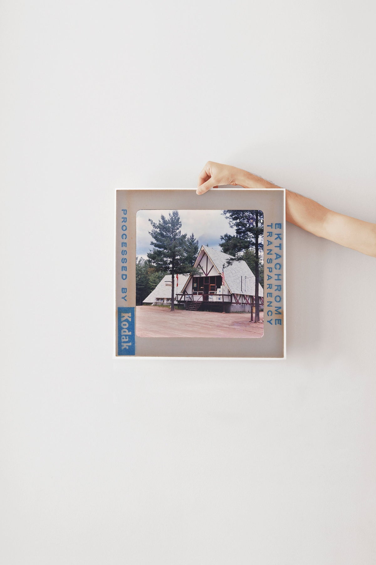 A hand is holding a framed photograph of a building with a triangular roof, surrounded by trees.