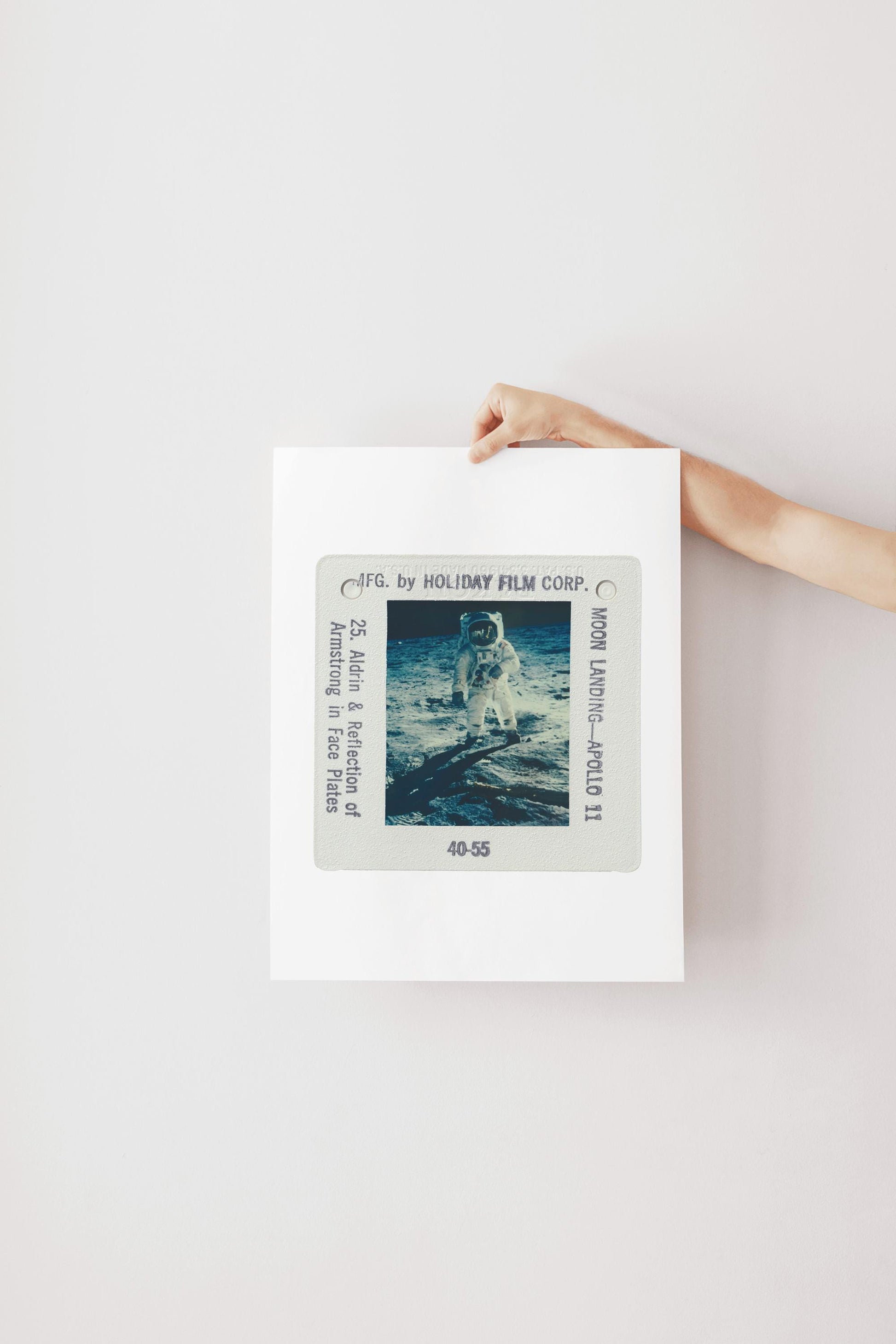 A person's hand is holding a framed photograph of an astronaut on the moon, with the frame displaying the text "F.G. by HOLIDAY FILM CORP. 40:55".