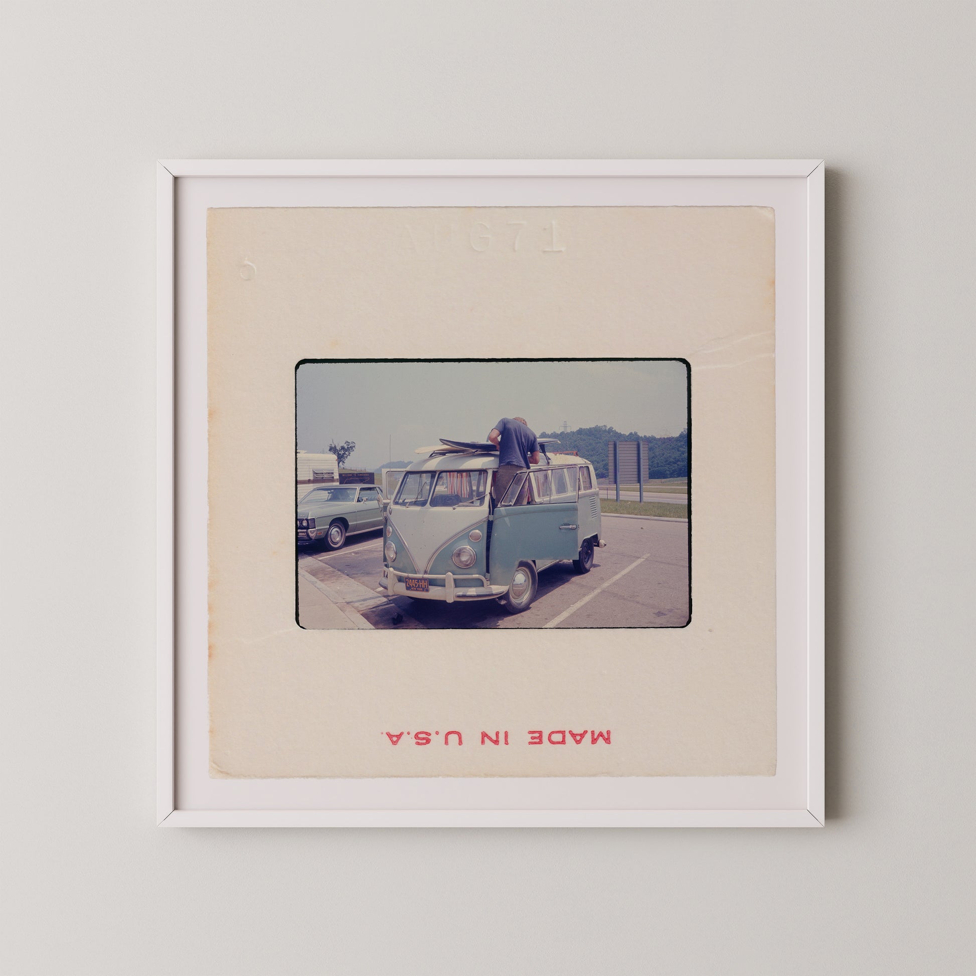 A vintage photograph of a blue Volkswagen van parked in a parking lot, with a person standing on the van's hood.