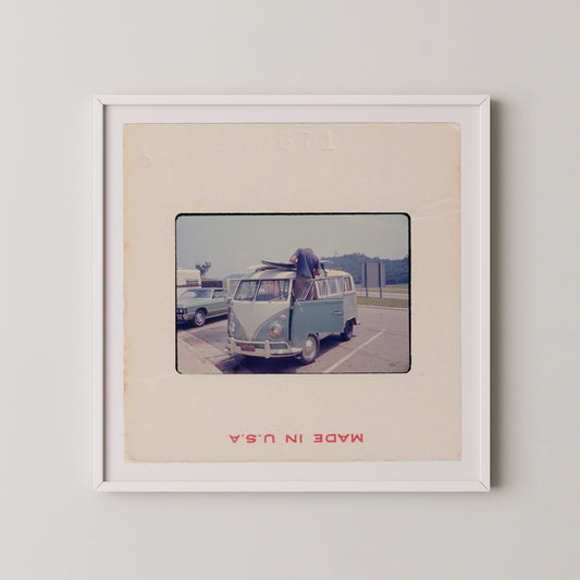 A vintage photograph of a blue Volkswagen van parked in a parking lot, with a person standing on the van's hood.