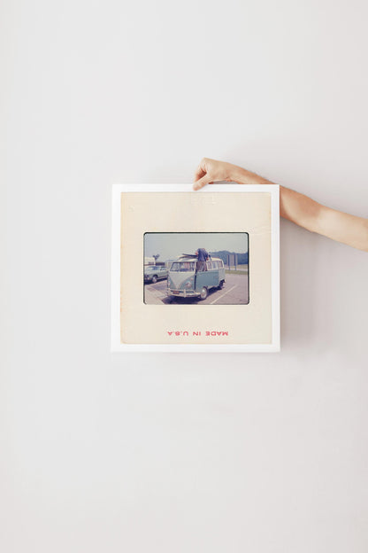 Vintage 1971 35mm slide photograph of a man loading two surfboards onto the roof rack of a pale blue Volkswagen Bus in a parking lot, with a "Welcome to Tennessee" highway sign and a vintage sedan visible in the background. Printed as a giclée fine art print on archival matte paper.
