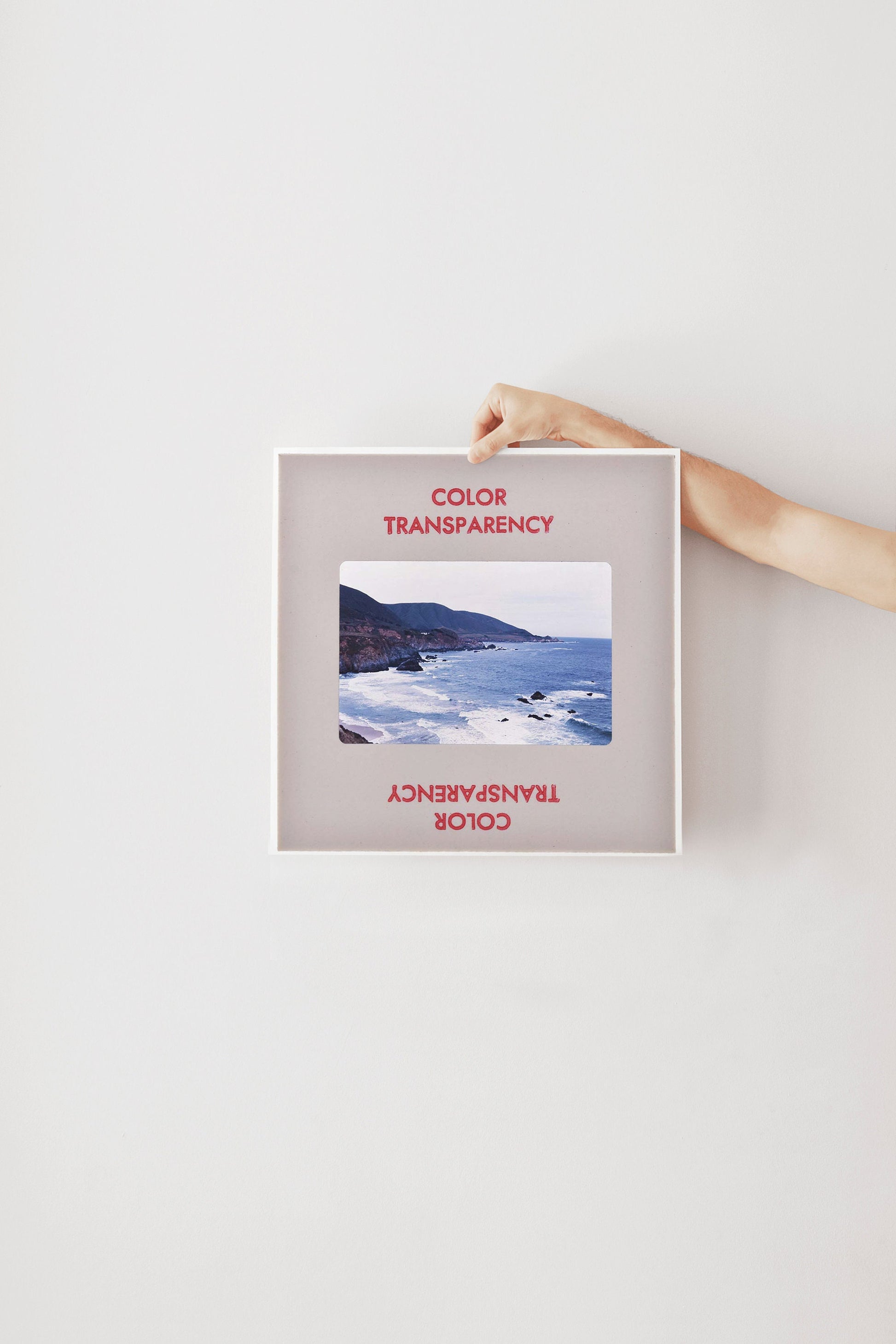 A person's hand is holding a framed photograph of a coastal landscape, with the text "COLOR TRANSPARENCY" and "COLOR" visible on the frame.