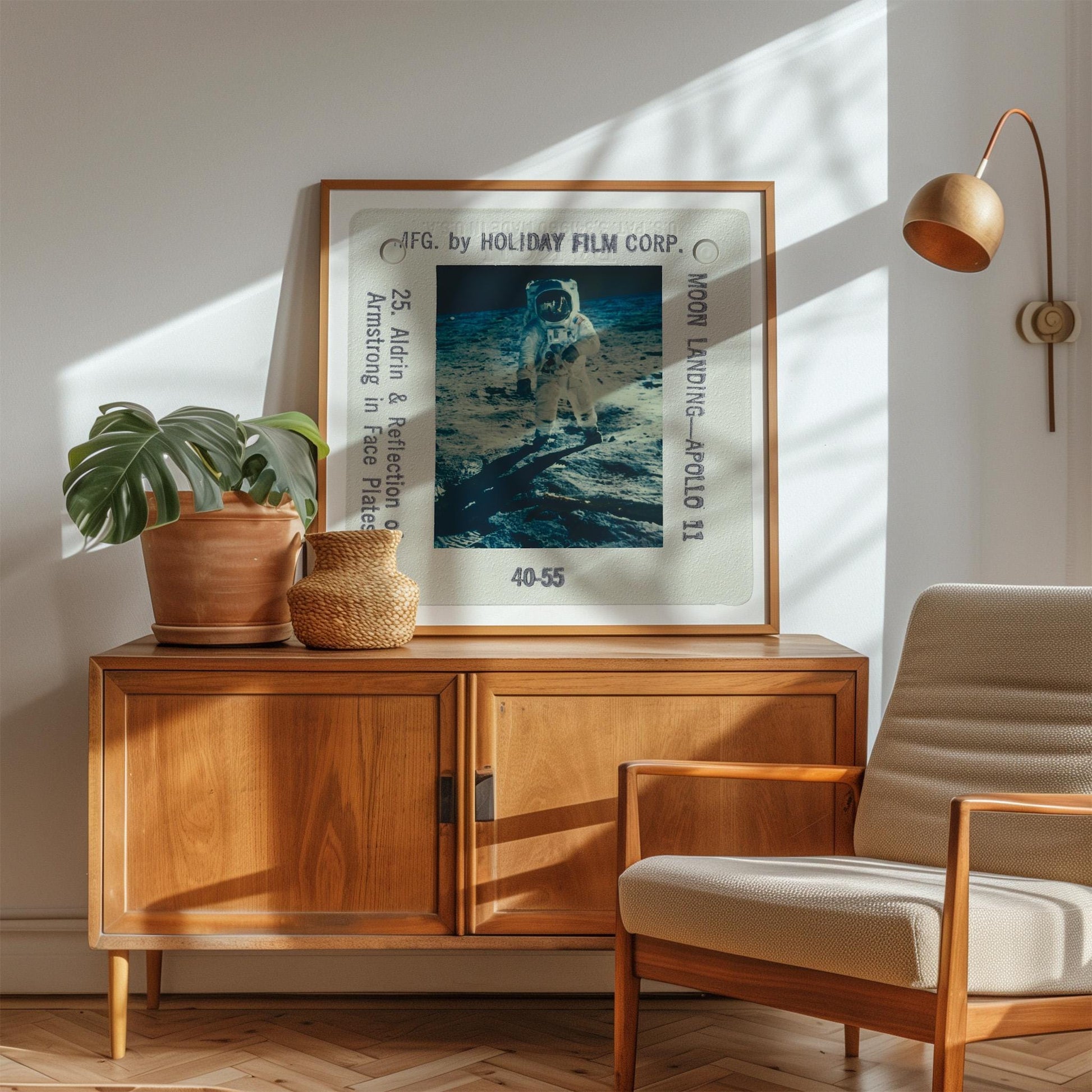 a framed photograph of an astronaut on the moon, displayed on a wooden cabinet or dresser. The photograph is framed by a white border and has the text "F.G. by HOLIDAY FILM CORP. Moon Landing - Apollo 11" printed on it. The cabinet has a wooden finish and a woven basket on top. In the background, there is a beige armchair and a potted plant.