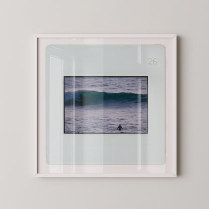 Vintage slide photograph of two surfers in the ocean — one riding a glassy teal-green wave in the background and one waiting chest-deep in silver-grey water in the foreground, shot with a telephoto lens from shore. Printed as a giclée fine art print on archival matte paper.