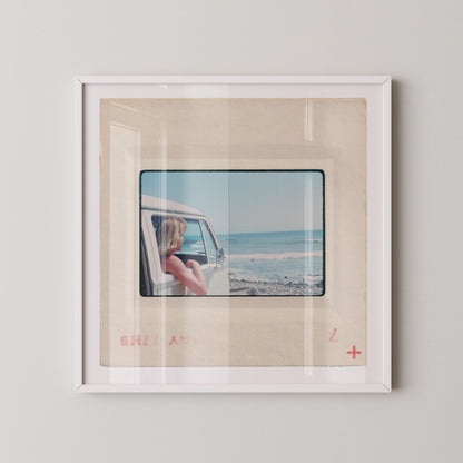 Vintage 1977 Kodachrome slide photograph of a blonde child leaning out the window of a white Volkswagen Microbus, looking toward the ocean along the California coast. Rocky shoreline and gentle surf visible in the background. Printed as a giclée fine art print on archival matte paper.